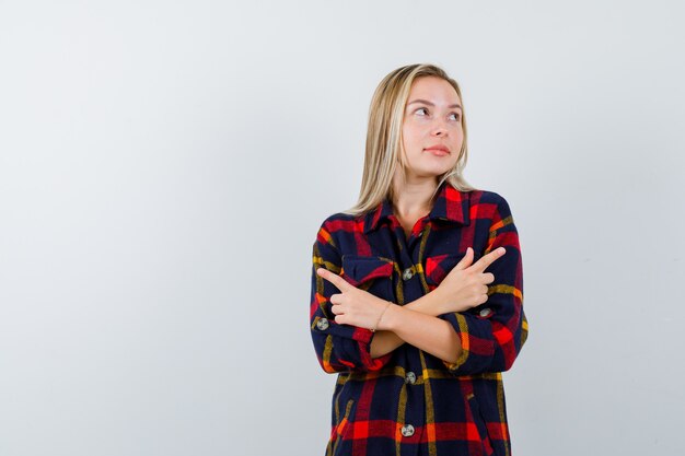 Retrato de señorita apuntando a ambos lados en camisa a cuadros y mirando vacilante vista frontal