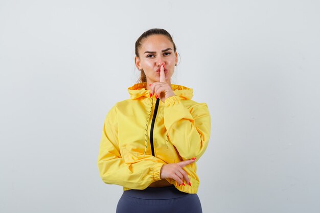 Retrato de señorita apuntando hacia el lado derecho, mostrando gesto de silencio en chaqueta amarilla y mirando confiada vista frontal