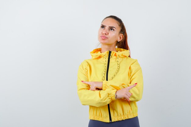 Retrato de señorita apuntando hacia los lados izquierdo y derecho, haciendo pucheros con los labios en una chaqueta amarilla y mirando confiada vista frontal