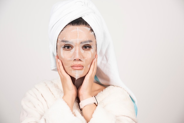 Retrato de señorita en bata de baño y toalla en la cabeza mientras toca su rostro con mascarilla.