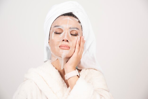 Foto gratuita retrato de señorita en bata de baño y toalla en la cabeza mientras toca su rostro con mascarilla.