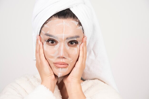 Retrato de señorita en bata de baño y toalla en la cabeza mientras toca su rostro con mascarilla.