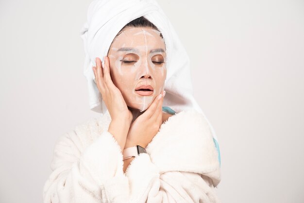 Retrato de señorita en bata de baño y toalla en la cabeza mientras toca su rostro con mascarilla