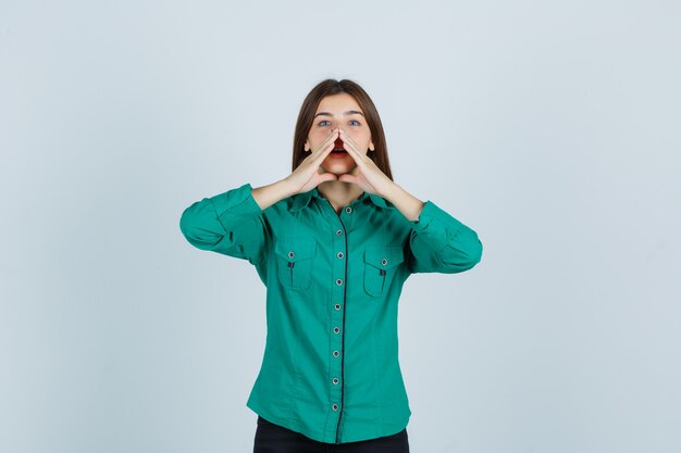 Retrato de señorita cogidos de la mano cerca de la boca mientras dice un secreto en camisa verde y mira curiosa vista frontal