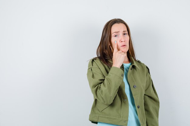 Retrato de señorita con el dedo en la mejilla en camiseta, chaqueta y mirando ofendido vista frontal