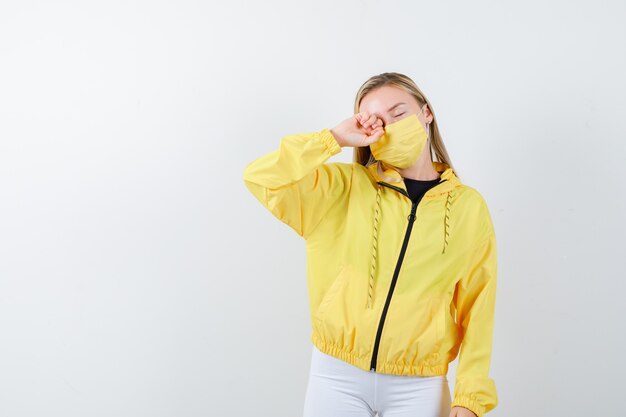 Retrato de señorita frotándose los ojos en chaqueta, pantalón, máscara y mirando soñoliento vista frontal