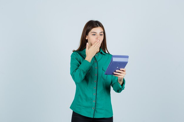 Retrato de señorita mirando calculadora mientras sostiene la mano en la boca en camisa verde y mirando sorprendido vista frontal