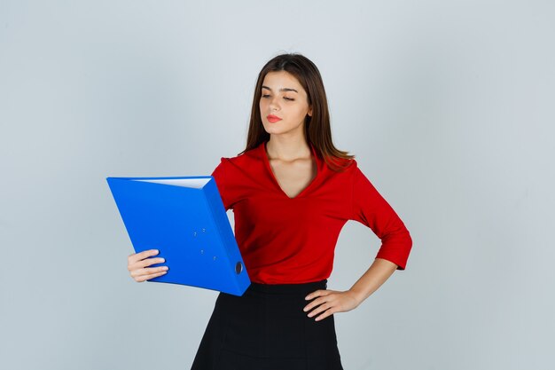 Retrato de señorita mirando carpeta mientras mantiene la mano en la cadera en blusa roja