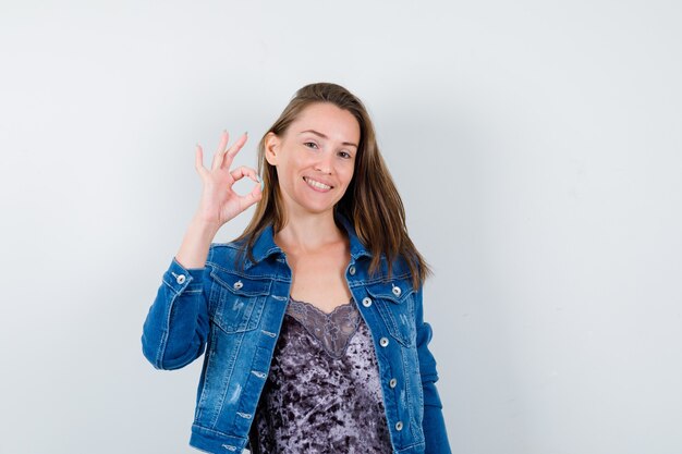 Retrato de señorita mostrando gesto ok en blusa, chaqueta de mezclilla y mirando confiado vista frontal