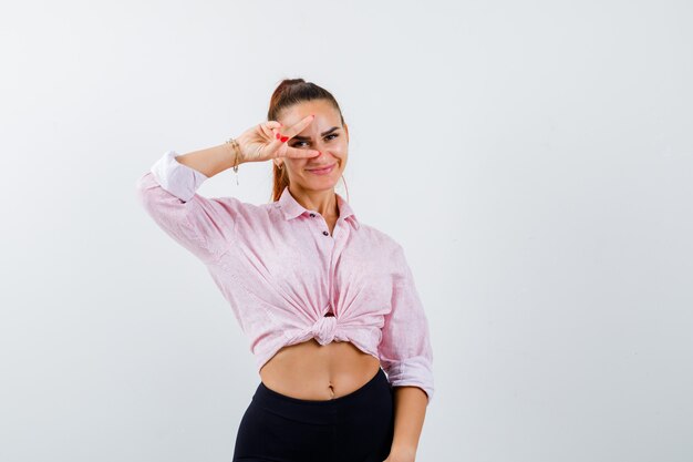 Retrato de señorita mostrando el signo V en el ojo en camisa, pantalones y mirando alegre vista frontal