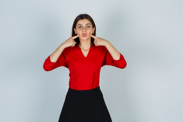 Retrato de señorita presionando los dedos en las mejillas en blusa roja