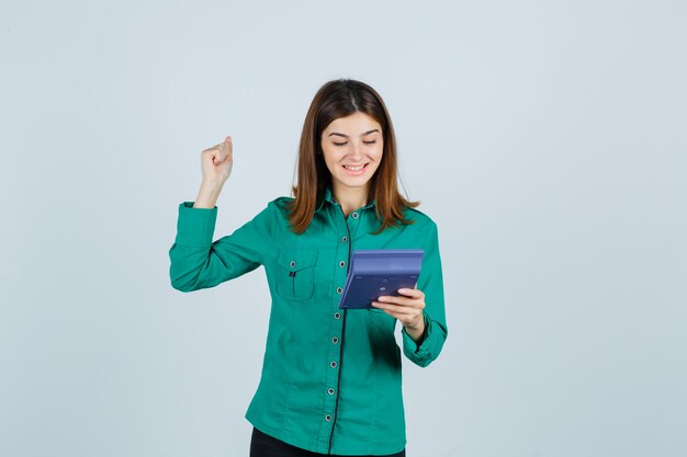 Retrato de señorita sosteniendo la calculadora, mostrando el gesto de ganador en camisa verde y mirando dichosa vista frontal