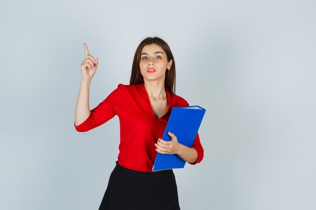 Retrato de señorita sosteniendo la carpeta mientras apunta hacia arriba en blusa roja