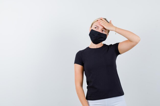 Retrato de señorita sosteniendo la mano en la frente en camiseta, pantalón, máscara médica