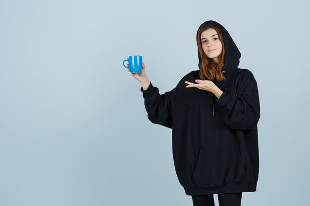 Retrato de señorita sosteniendo una taza mientras finge mostrar algo en una sudadera con capucha de gran tamaño, pantalones y mirando confiada vista frontal