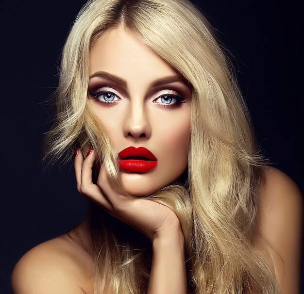 Retrato sensual glamour de hermosa mujer rubia modelo dama con maquillaje brillante y labios rojos tocando su rostro, con cabello rizado saludable sobre fondo negro