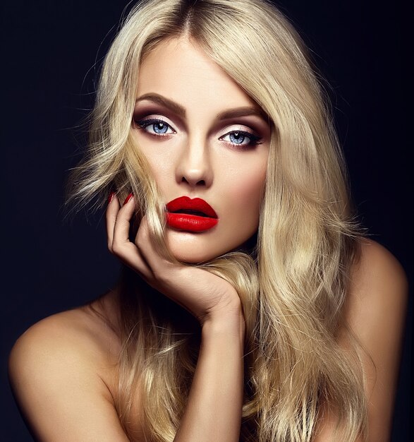 Retrato sensual glamour de hermosa mujer rubia modelo dama con maquillaje brillante y labios rojos tocando su rostro, con cabello rizado saludable sobre fondo negro