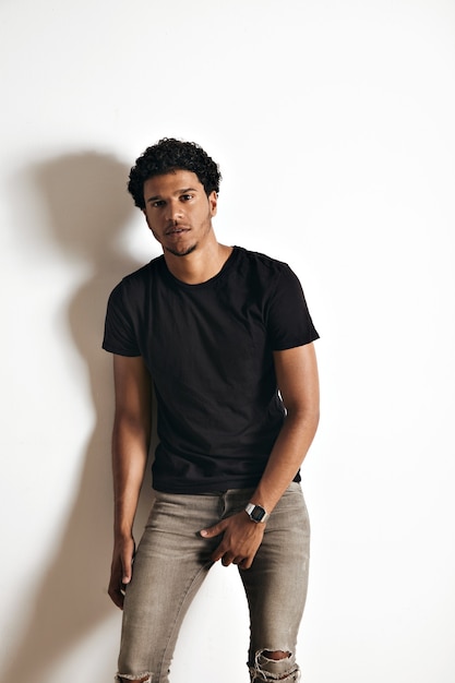 Retrato sensual de un modelo afroamericano joven musculoso guapo en jeans grises delgados y camiseta de algodón negro liso de pie junto a una pared blanca.