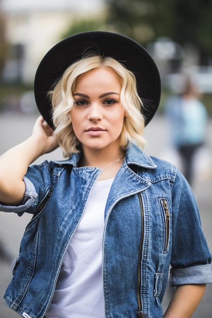 Retrato de seria chica rubia en el centro de la ciudad vestida con jeans suite y camiseta blanca y sombrero negro