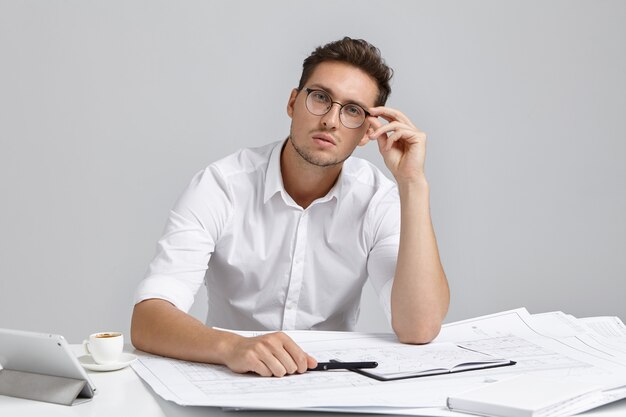 Retrato de serio arquitecto masculino confiado trabaja en planos, viste camisa blanca formal y gafas redondeadas