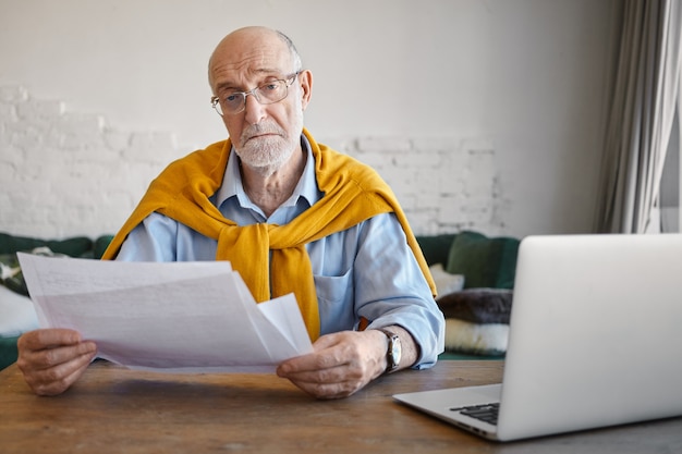 Retrato de serio empresario anciano exitoso con elegante atuendo y accesorios revisando documentos financieros en sus manos, mientras trabaja en la oficina moderna, usando un dispositivo electrónico