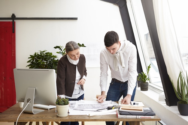 Retrato de serios diseñadores profesionales creativos, hombre joven y mujer mayor que trabaja en el proyecto, de pie en el escritorio de oficina, creando diseños de interiores de casas residenciales y propiedades comerciales