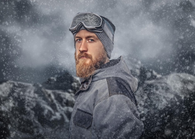 Retrato de un snowboarder pelirrojo con barba completa en un sombrero de invierno y gafas protectoras vestidas con un abrigo de snowboard posando contra el fondo de las montañas.