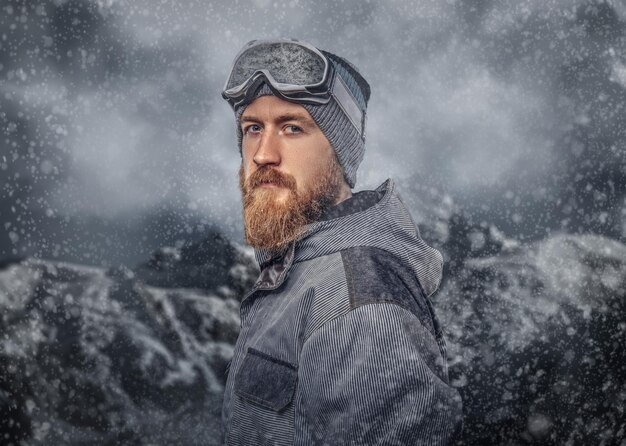 Retrato de un snowboarder pelirrojo con barba completa en un sombrero de invierno y gafas protectoras vestidas con un abrigo de snowboard posando contra el fondo de las montañas.