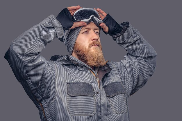 Retrato de un snowboarder pelirrojo con barba completa en un sombrero de invierno y gafas protectoras vestidas con un abrigo de snowboard posando en un estudio, mirando hacia otro lado. Aislado en el fondo gris.