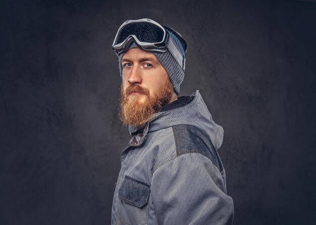 Retrato de un snowboarder pelirrojo con barba completa en un sombrero de invierno y gafas protectoras vestidas con un abrigo de snowboard posando en un estudio, mirando hacia otro lado. Aislado en un fondo de textura oscura.