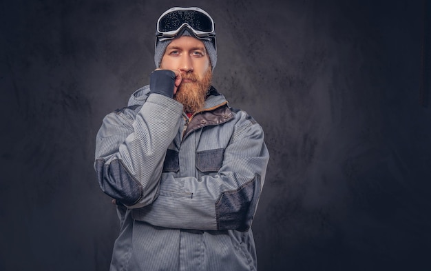 Retrato de un snowboarder pelirrojo pensativo con barba completa en un sombrero de invierno y gafas protectoras vestidas con un abrigo de snowboard posando en un estudio, mirando hacia otro lado. Aislado en un backgrou de textura oscura