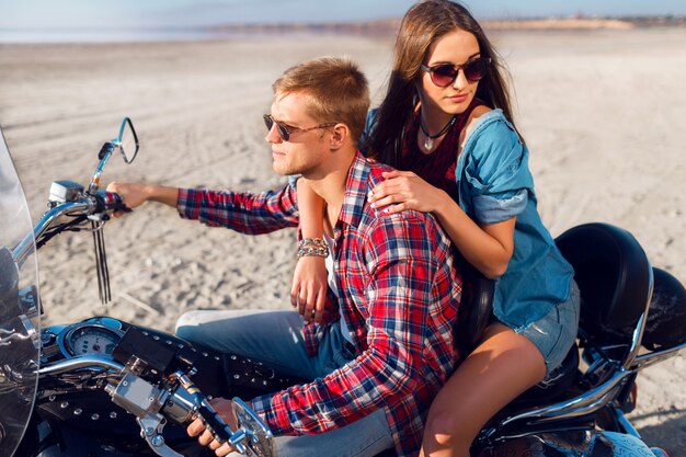 Retrato soleado de estilo de vida de jóvenes jinetes de pareja sentados juntos en la playa de arena en moto - concepto de viaje. Dos personas y bicicleta. Moda mujer y hombre abrazando y sonriendo.