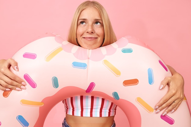 Retrato de soñadora linda rubia adolescente en ropa de verano posando aislada con anillo de natación rosa en forma de rosquilla, mirando hacia arriba, soñando con vacaciones en el mar