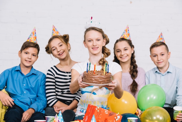 Retrato sonriente de una adolescente que se sienta con sus amigos que sostienen la torta de cumpleaños