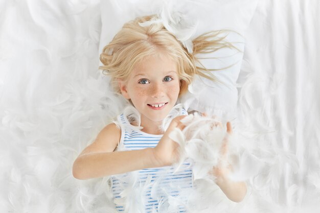 Retrato de sonriente alegre niña caucásica con cabello rubio y pecas jugando con plumas blancas mientras está acostado en la cama, con expresión alegre y juguetona en su rostro bastante infantil