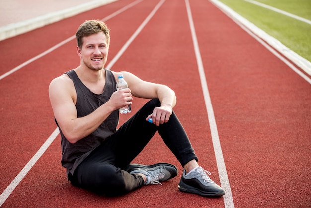 Retrato sonriente de un atleta masculino sentado en la pista de carreras con una botella de agua en la mano