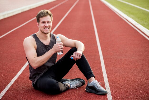 Retrato sonriente de un atleta masculino sentado en la pista de carreras con una botella de agua en la mano