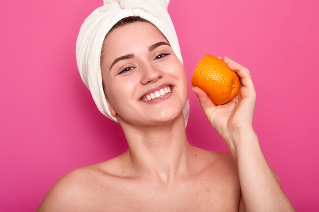 Retrato de sonriente atractiva mujer medio desnuda, la señora sostiene rodajas de naranja en su rostro y mira a la cámara aislada en rosa. Linda mujer con toalla en la cabeza cuida la piel en el baño.