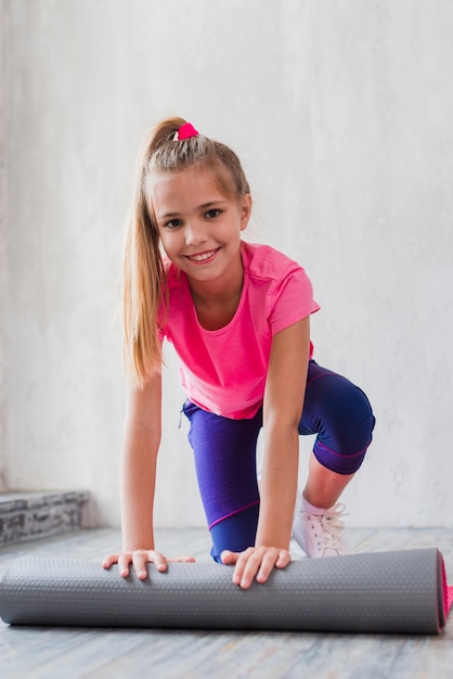 Retrato sonriente de una chica rubia que rueda la estera del ejercicio delante de la pared