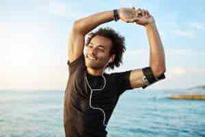 Foto gratuita retrato de un sonriente deportista afroamericano estirando sus musculosos brazos antes de entrenar junto al mar, usando la aplicación de música en su teléfono inteligente.