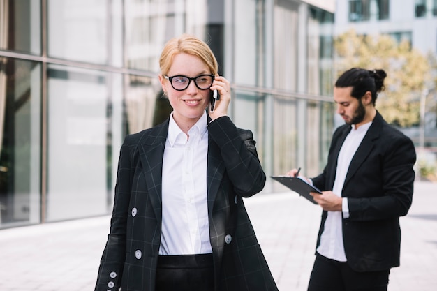 Foto gratuita retrato sonriente de una empresaria joven que habla en el teléfono celular con la escritura del hombre en el tablero en el fondo