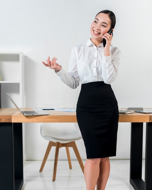 Retrato sonriente de una empresaria que se coloca delante del escritorio que habla en gesticular del teléfono móvil