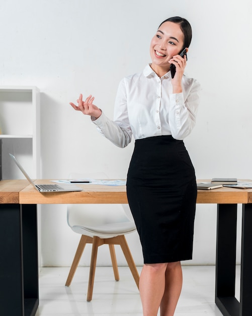 Retrato sonriente de una empresaria que se coloca delante del escritorio que habla en gesticular del teléfono móvil