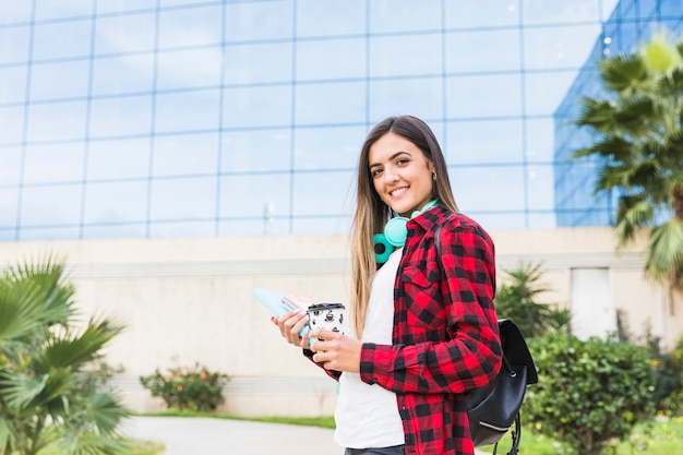 Retrato sonriente de un estudiante joven que sostiene los libros y la taza de café para llevar que se coloca delante del edificio de la universidad