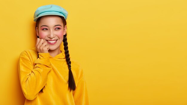 Retrato de sonriente hermosa niña de cabello oscuro con dos coletas, tiene maquillaje de color rosa brillante, viste elegante sombrero y suéter de pana con capucha