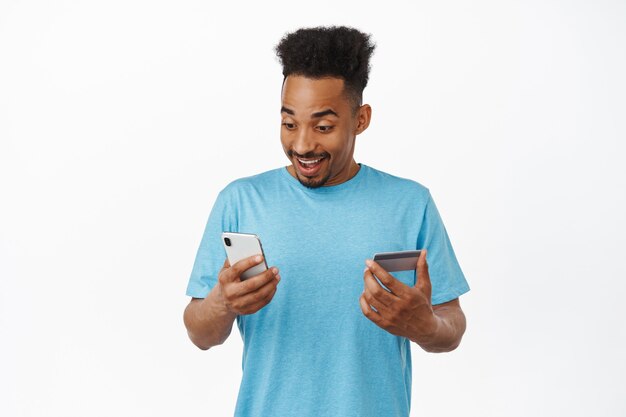 Retrato de sonriente hombre afroamericano con teléfono inteligente y tarjeta de crédito, pagando en línea, comprando en la aplicación, de pie en camiseta azul sobre blanco