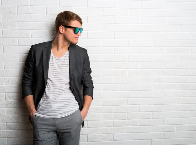 Retrato de sonriente hombre elegante en gafas de sol de pie contra la pared de ladrillo en la oficina moderna.