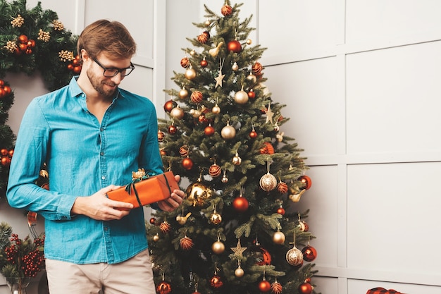 Retrato de sonriente hombre guapo con regalo. Hombre barbudo sexy posando cerca del árbol de Navidad con presente.