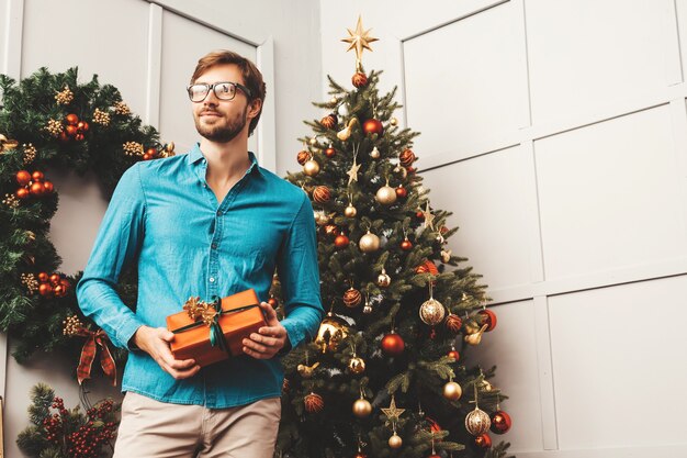 Retrato de sonriente hombre guapo con regalo. Hombre barbudo sexy posando cerca del árbol de Navidad con presente.