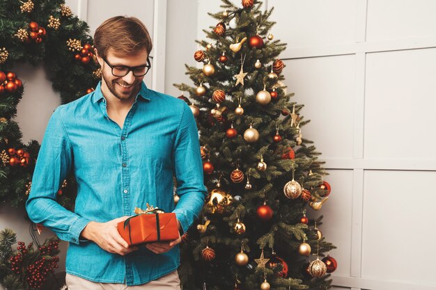 Retrato de sonriente hombre guapo con regalo. Hombre barbudo sexy posando cerca del árbol de Navidad con presente.